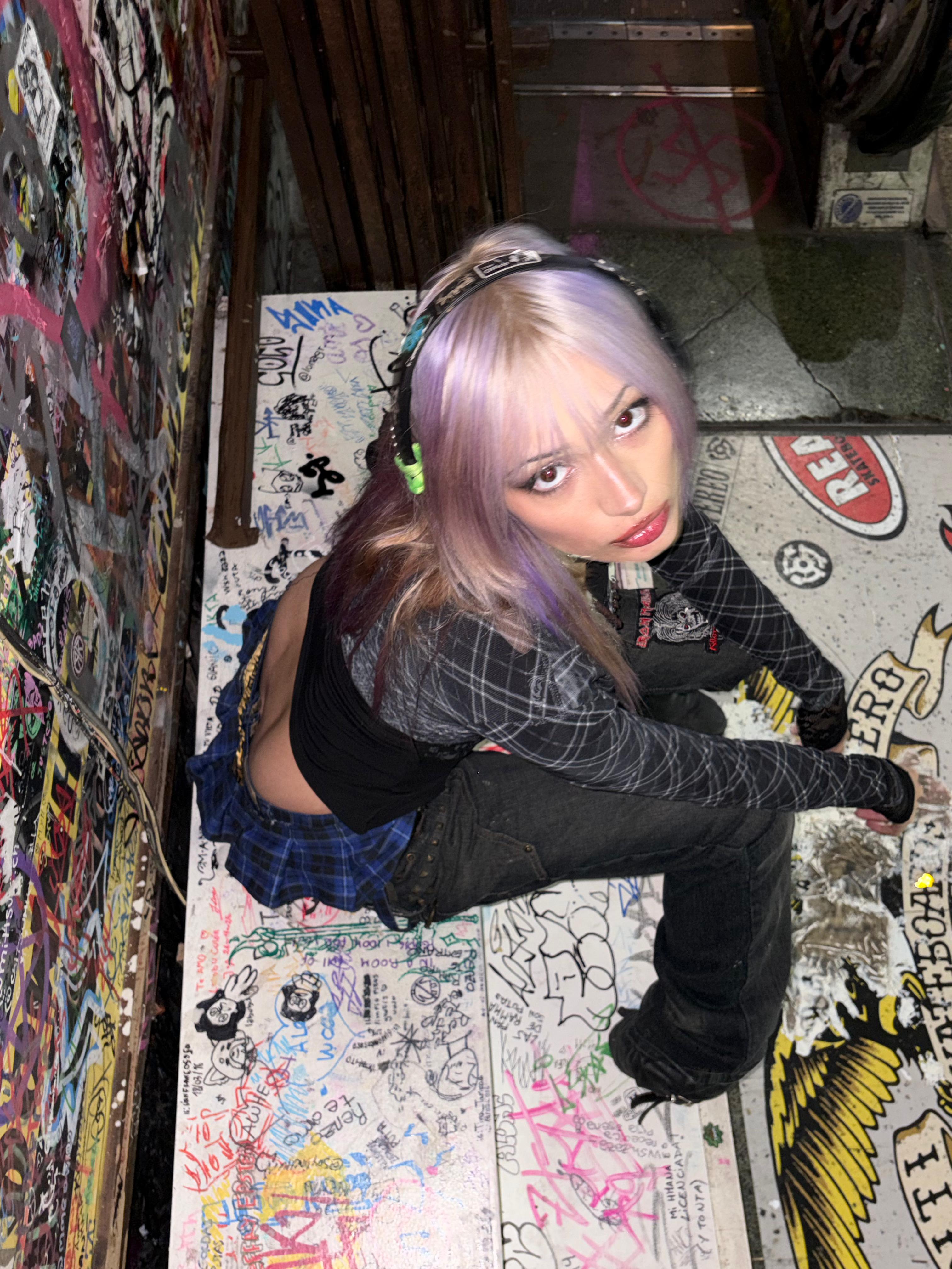 Elena crouched in a graffiti-covered skate shop stairwell in a spiderweb top and plaid skirt
