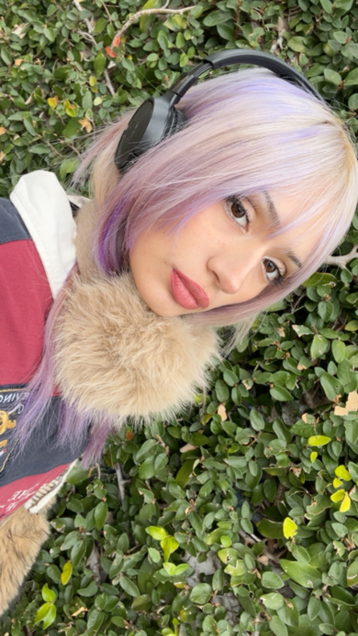 Elena outdoors against a green hedge wearing headphones and a red and white varsity jacket with fur collar
